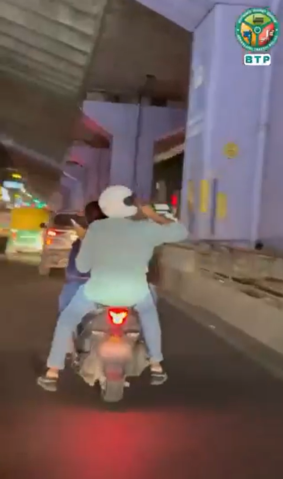 Rear view of two scooter riders on a city road beneath a flyover, with the Bengaluru Traffic Police logo visible