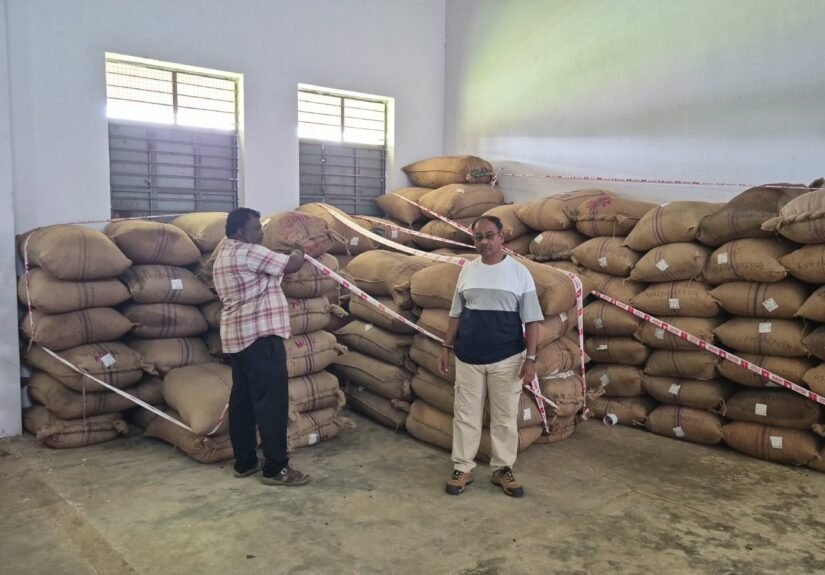 Seized arecanut bags stored in a godown in Kolar, with two men standing beside the consignment