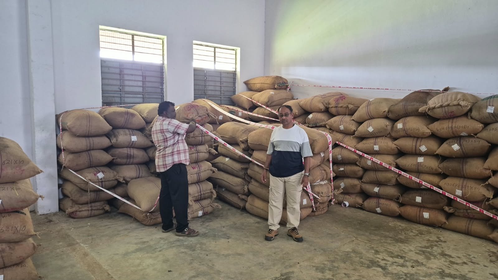 Seized arecanut bags stored in a godown in Kolar, with two men standing beside the consignment